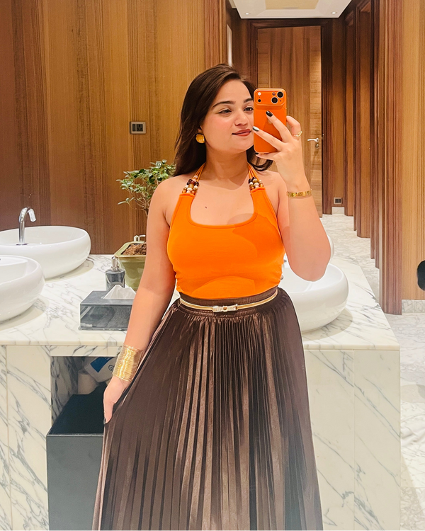 A woman takes a mirror selfie in a luxury bathroom, wearing a bright orange sleeveless top and a pleated dark brown skirt.