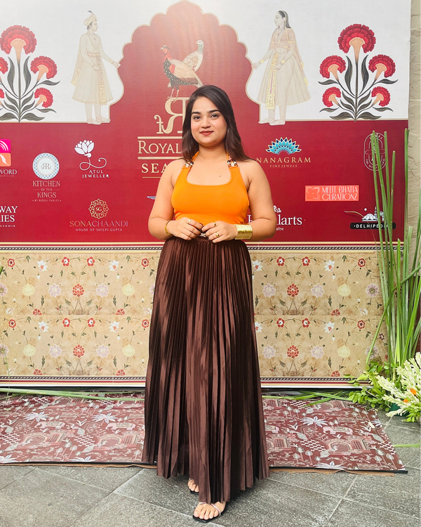 A woman stands barefoot in front of a decorative event backdrop, wearing a sleeveless mustard yellow top and a long pleated dark brown skirt.